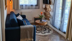 A modern living room with a blue sofa, a round wooden coffee table, a hanging light fixture, a mounted TV, and a large window with curtains. - Gîtes de France® - At the crossroads (BERNEVAL LE GRAND)