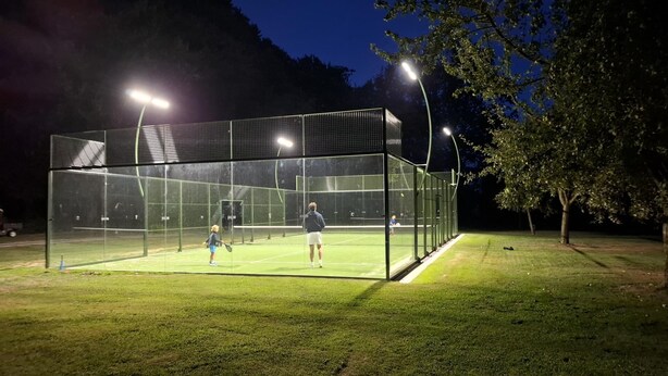Een binnentennisbaan met net, omgeven door een metalen hekwerk en verlicht met hangende lampen.