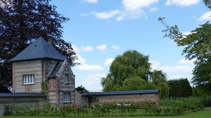 Een twee verdiepingen tellend gebouw van steen met een puntige gevel, omringd door een verzorgd gazon en bloeiende struiken.