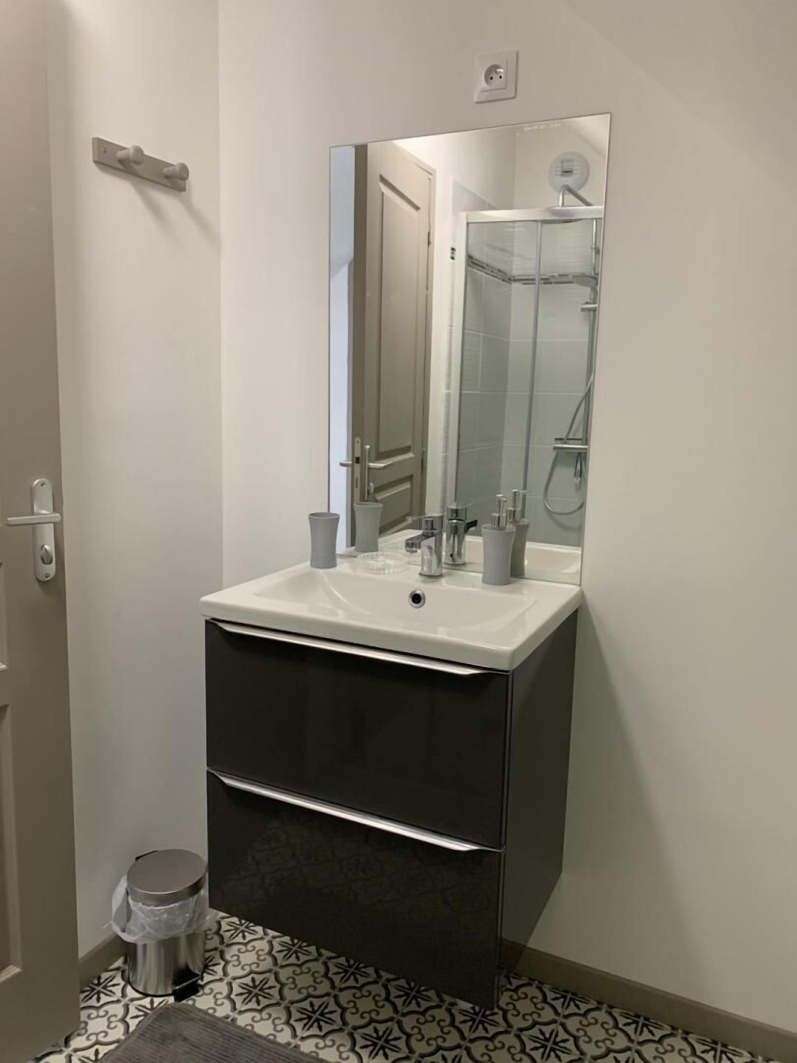 Une salle de bain avec un lavabo blanc, un miroir et une douche. On y trouve une poubelle et un carrelage décoré.