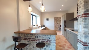 A kitchen with a central island, two bar stools, hanging pendant lights, a stone wall, and a brick backsplash.