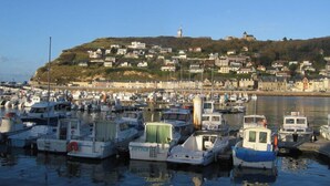 Una marina con diverse imbarcazioni ormeggiate, una collina con edifici e un faro sullo sfondo.
