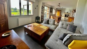 A living room with a sofa, a wooden coffee table, and a dining table. There are large windows providing a view of the outdoors. - Gîtes de France® - La Demeure des Vents (LE TILLEUL)