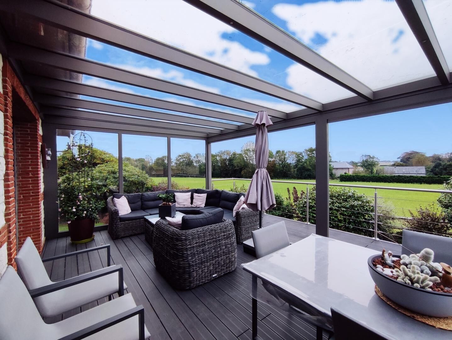 A covered outdoor seating area with a glass roof, a cushioned sofa, a table, and a cactus display.