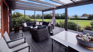 A covered outdoor seating area with a glass roof, a cushioned sofa, a table, and a cactus display. - Gîtes de France® - La Demeure des Vents (LE TILLEUL)