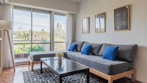 Un salon moderne avec un canapé gris, des coussins bleus, une table basse en verre et une vue sur la verdure et les bâtiments, grâce à de grandes fenêtres.