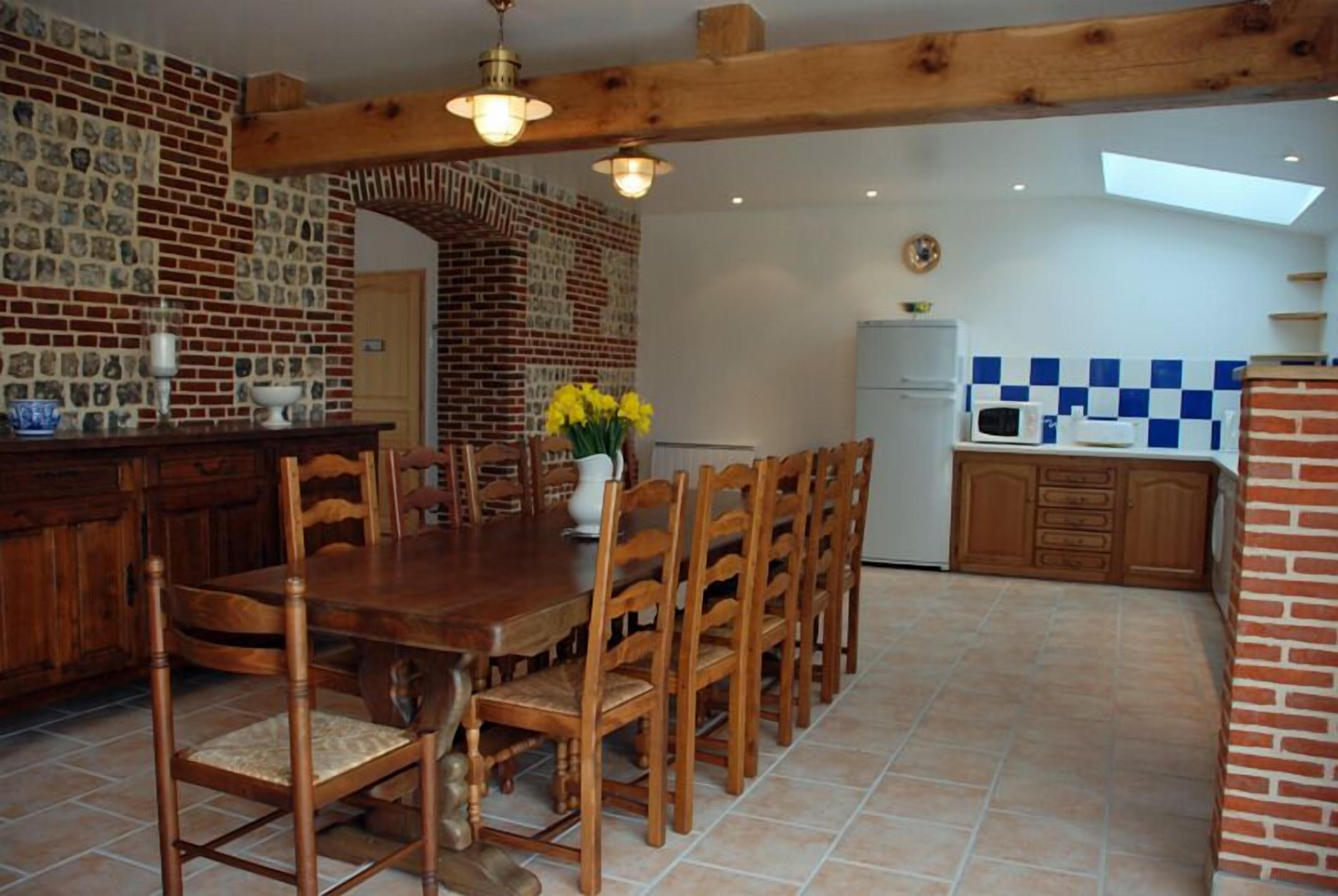 Uma área de refeições com mesa e cadeiras de madeira, parede de tijolos com um relógio, e uma cozinha com azulejos quadriculados azuis e brancos.