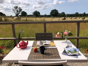 A table with a red teapot, a black mug, a book, and a bouquet of flowers on a deck with a view of hay bales and trees. - Gîtes de France® - Le Petit Giteopré (BEAUBEC LA ROSIERE)