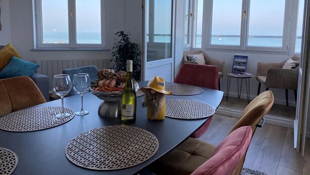 A dining area with a table set for two, a bottle of wine, and a decorative hat. The room has large windows with a view of the sea.