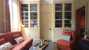 A cozy living room with a red sofa, a blue armchair, and a wooden coffee table with decorative items. There are two large glass-fronted cabinets with shelves, and a small cabinet with a vase on the right. - Gîtes de France® - Bayen's Family (FECAMP)