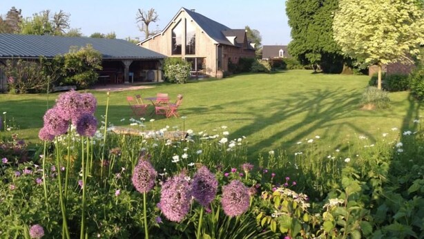 Ein Haus mit geneigtem Dach, einer überdachten Veranda und einem gepflegten Rasen mit Blumen und einem Baum.