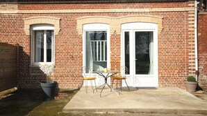 Una casa de ladrillo con dos ventanas enmarcadas en blanco y una puerta, un patio de concreto con una mesa y sillas, y una planta en maceta.