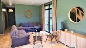 Ein Wohnzimmer mit einer Couch, zwei Sesseln, einem kleinen Tisch und einem Korbsessel. An der Wand befindet sich ein Spiegel, ein Kranz und ein Fenster mit Vorhängen.