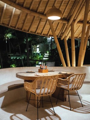 An outdoor dining area with a round table and four wicker chairs, surrounded by a bamboo ceiling structure. - Neano Escape (Manggis)