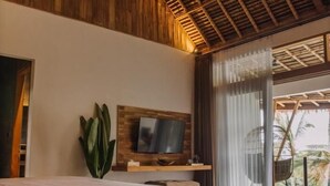 Quarto com teto de madeira, uma cama, uma TV em uma estante de madeira e vista para palmeiras através de grandes janelas.