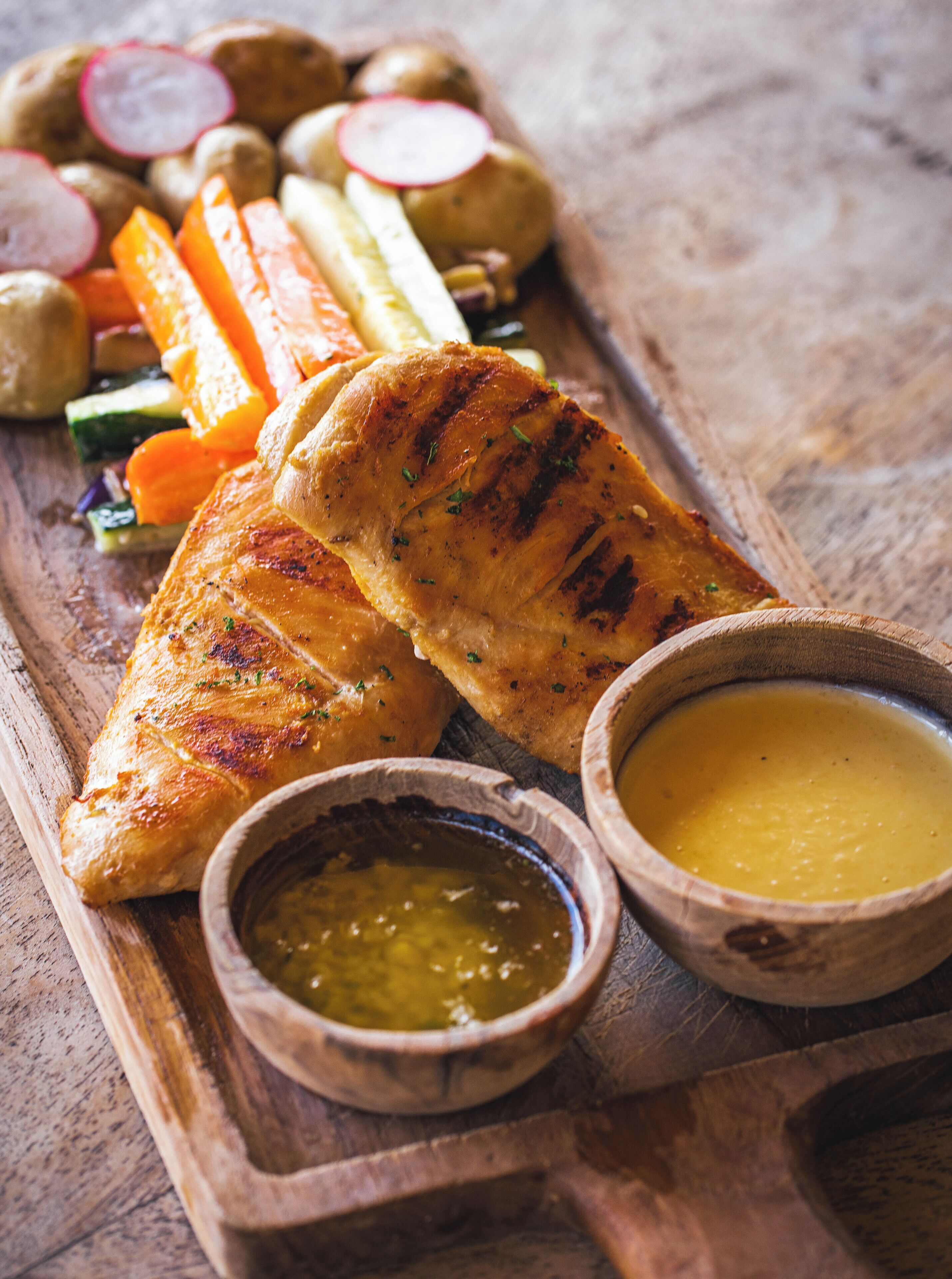 A wooden serving tray with grilled chicken, roasted vegetables, and two small bowls of sauce.