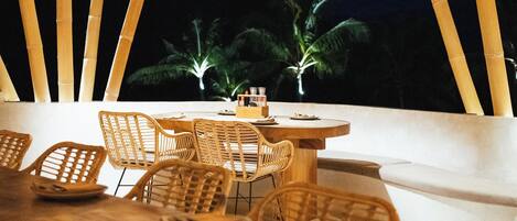 An outdoor dining area with bamboo ceiling and chairs, a table with plates and cutlery, and a view of palm trees at night.