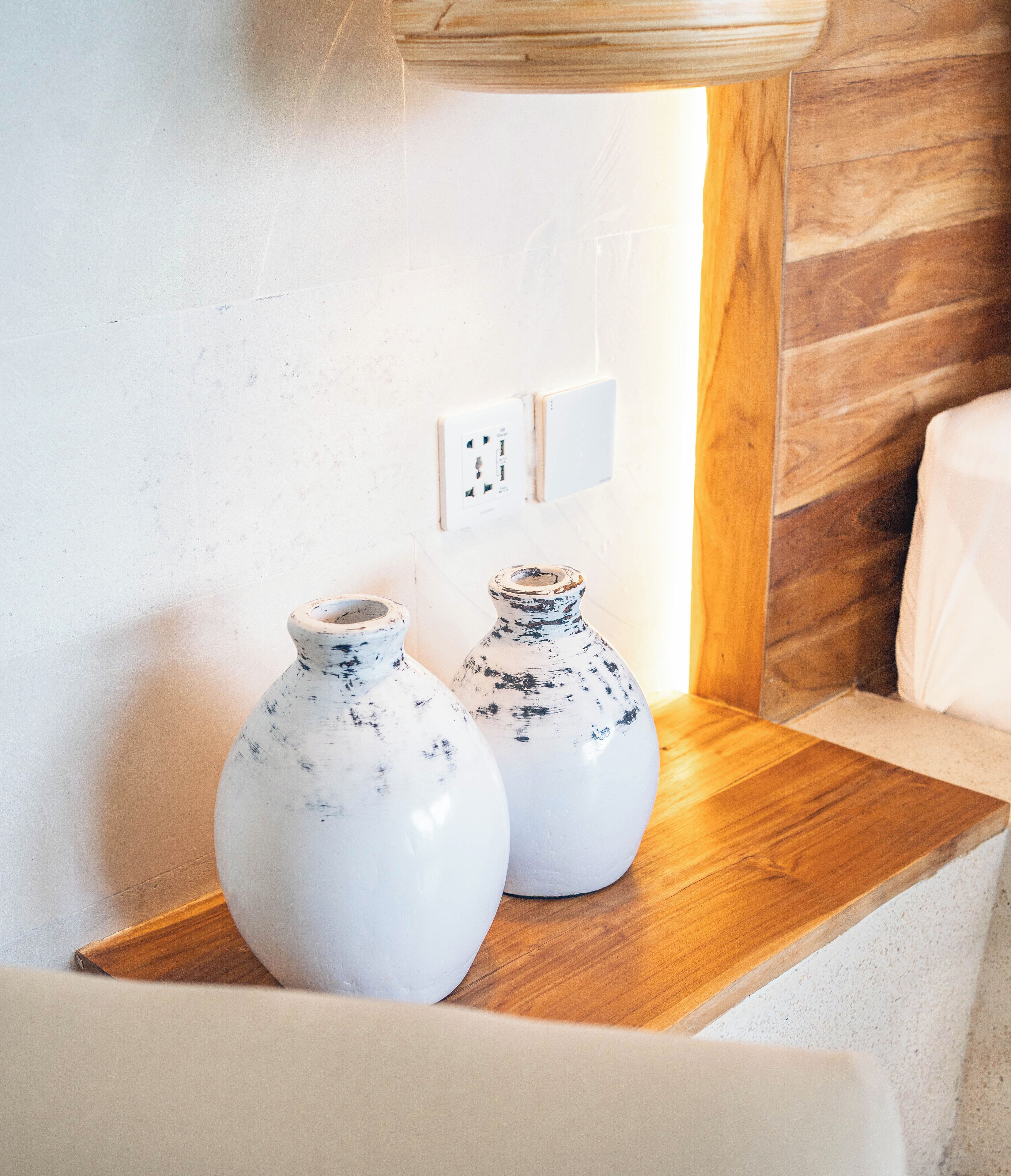 Two white ceramic vases with blue speckles on a wooden shelf in a room with a white wall and a wooden panel.