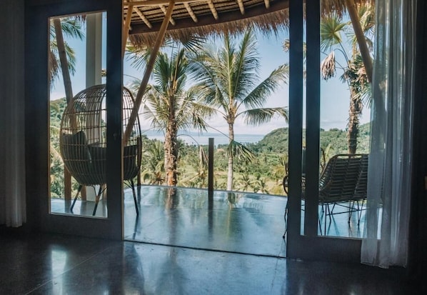 A room with a large glass door leading to a balcony with a view of palm trees and hills. - Neano Escape (Manggis)