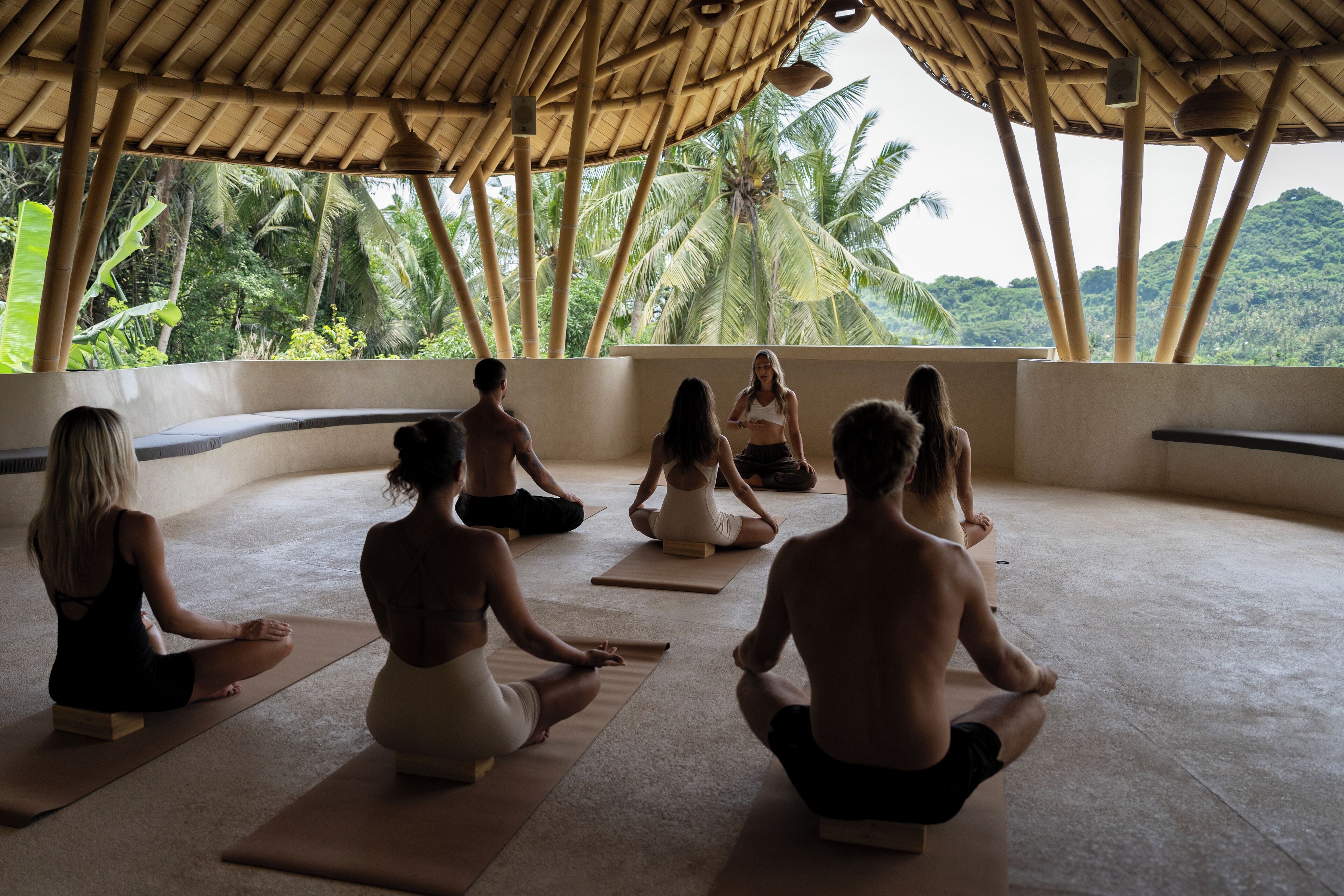 A group of people meditating in a room with a view of palm trees.