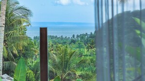 Vista da janela com paisagem tropical, palmeiras e um corpo de água.