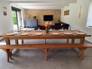 Un comedor con una larga mesa de madera dispuesta para seis comensales, un aparador azul, un televisor montado en una pared de piedra y un sofá al fondo.