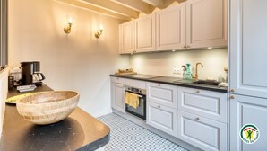 A modern kitchen with white cabinets, a black countertop, and a wooden bowl. There are two wall-mounted lights and a coffee machine on the counter. - Gîtes de France® - Chez Mlle Rose (VEULES LES ROSES)