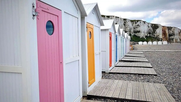 Eine Reihe von Strandhäusern mit bunten Türen, Holzstegen und einer felsigen Klippe im Hintergrund.