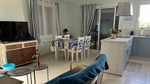 A living room with a sofa, a TV, a dining table with chairs, a kitchen area, and a sliding glass door. - Gîtes de France® - Le Gîte des 2 Falaises (EU)