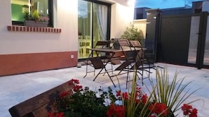 An outdoor patio with a table and chairs, a small garden area with red flowers, and a glass door leading to a room. - Gîtes de France® - Le Gîte des 2 Falaises (EU)