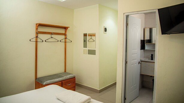 Quarto com cama, um banquinho, um cabideiro de madeira, um espelho e uma porta para o banheiro.
