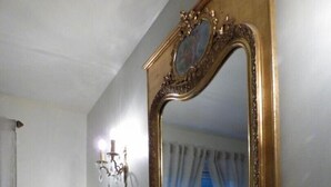 A bathroom with a large mirror, a vanity with a marble countertop, a small table with a lamp, and a wall-mounted light fixture. - Gîtes de France® - Le Clos des Camélias (VEULETTES SUR MER)