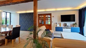 A modern living room with a wooden ceiling beam, a dining area with a table and chairs, a sofa, a TV, and a bookshelf.