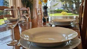 Mesa de jantar posta com pratos, copos e talheres, vista pela janela de um jardim e casas.