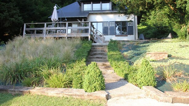 A house with a covered porch, a staircase leading to the entrance, and a garden with various plants and a wooden deck.