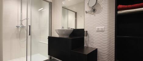 Un baño moderno con cabina de ducha de vidrio, un tocador negro y un lavamanos tipo recipiente, y una pared con textura.