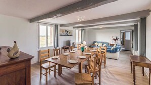A spacious dining area with a wooden table set for six, flanked by wooden chairs. A rustic wooden cabinet is visible on the left, and a sofa with a blue blanket is in the background. - Gîtes de France® - Gîte d'Eclehêtre (FOUCARMONT)