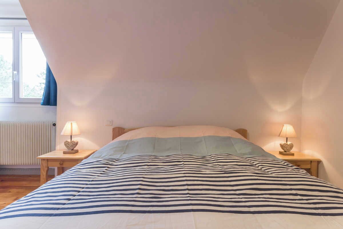 Une chambre avec un plafond en pente, un lit avec une literie à rayures, deux tables de chevet avec des lampes et une fenêtre avec des rideaux.
