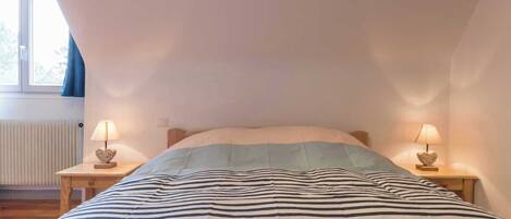 A bedroom with a sloped ceiling, a bed with striped bedding, two bedside tables with lamps, and a window with curtains.