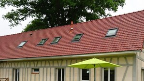 A two-story building with a red-tiled roof, a balcony with a yellow umbrella, and a garden area with a table and chairs.