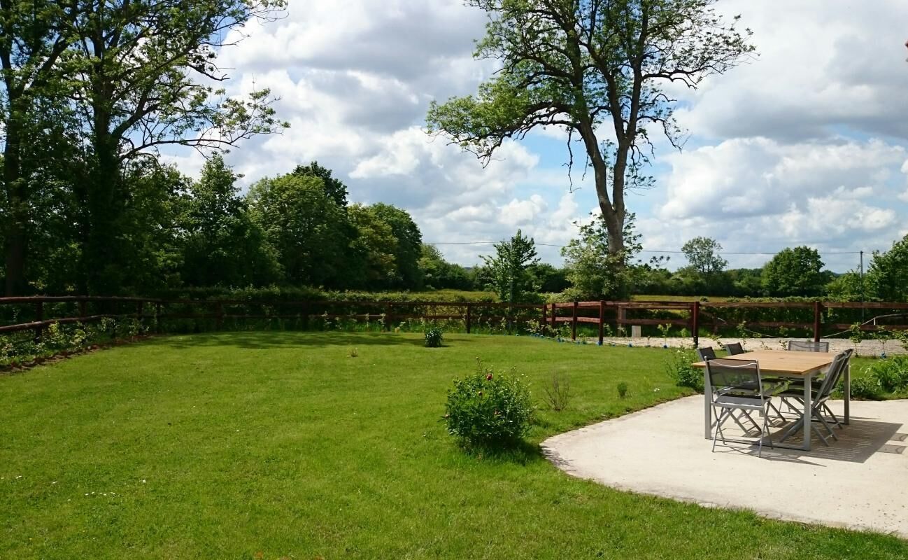 Een gazon met een houten tafel en stoelen, omgeven door bomen en een haag.