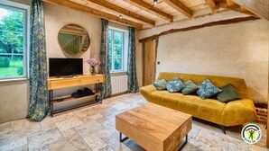 A cozy living room with a wooden ceiling, a yellow sofa, a wooden coffee table, a TV on a wooden stand, and patterned curtains. - Gîtes de France® - The Cottage (BOUDEVILLE)