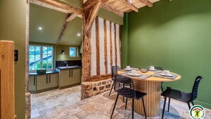 A rustic kitchen with stone walls and wooden beams, a dining area with a round table and black chairs, and a view of the outdoors through a window. - Gîtes de France® - The Cottage (BOUDEVILLE)