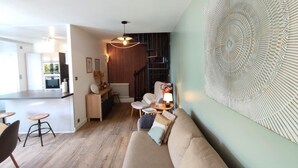 Una sala moderna con un sofá, un comedor con taburetes, una cocina al fondo y un elemento decorativo en la pared.