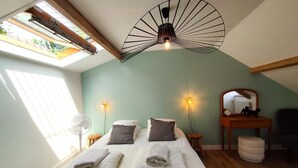 Un dormitorio con tragaluz, una cama con almohadas, una mesita de noche, un espejo y una lámpara fijada a la pared.