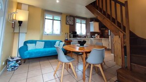 A cozy living room with a blue sofa, a wooden dining table with chairs, a kitchen area, and a staircase. - Gîtes de France® - Le Four à Chaux (CRIEL SUR MER)