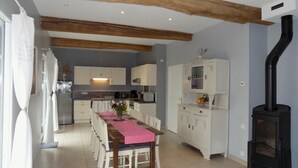 A kitchen with wooden beams, a dining area with a pink tablecloth, and a black wood stove. - Gîtes de France® - Le Neuf-Moulin (SAINT CRESPIN)