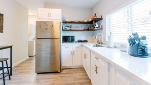 A modern kitchen with white cabinets, a stainless steel refrigerator, a microwave, and a sink. There are wooden shelves above the counter holding various kitchen items.
