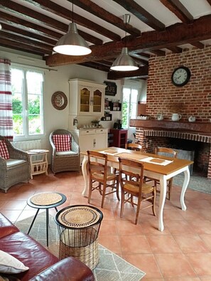 Un interior rústico con vigas de madera a la vista, una chimenea de ladrillo, una mesa de comedor con sillas, muebles de mimbre y una zona de cocina.