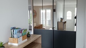 Un intérieur moderne avec un bureau en bois, des livres et une plante. Une porte coulissante donne accès à un balcon offrant une vue sur l’extérieur.